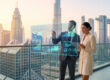 Un homme et une femme d'affaires analysent des données économiques sur un écran holographique devant la skyline de Dubaï.