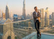 Un homme en costume pose sur un balcon surplombant les gratte-ciels de Dubaï au coucher du soleil.