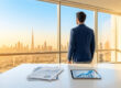 Un homme d'affaires regarde la skyline de Dubaï depuis un bureau avec des graphiques financiers sur la table.