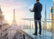 Un homme d'affaires sur un balcon surplombant Paris et Dubaï avec une tablette holographique.