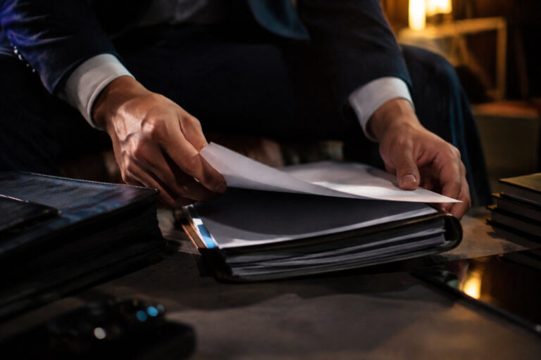 Gros plan sur les mains d'un homme en costume feuilletant des documents officiels sur un bureau sombre.