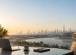 Un ordinateur portable, un café et un carnet sur une table avec une vue panoramique sur les gratte-ciels de Dubaï.