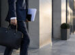 Un homme d'affaires en costume tenant une mallette et des documents devant un immeuble moderne.