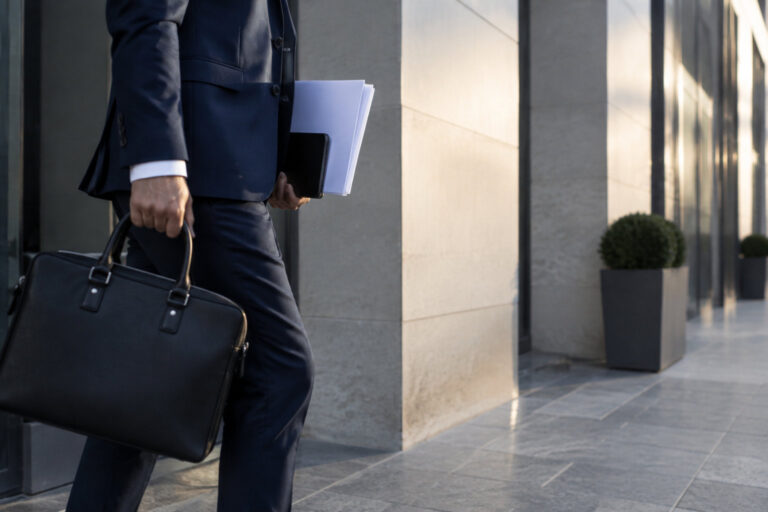 Un homme d'affaires en costume tenant une mallette et des documents devant un immeuble moderne.