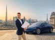 Un homme en costume tient des documents officiels devant une Tesla bleue, avec Dubaï et Paris en arrière-plan.