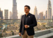 Un professionnel souriant en blazer noir devant la ligne d'horizon de Dubaï au coucher du soleil.